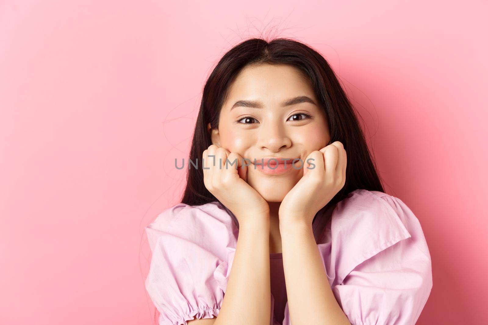 Close-up of excited asian girl listening with interest, smiling amused and looking at camera, standing against pink background by Benzoix