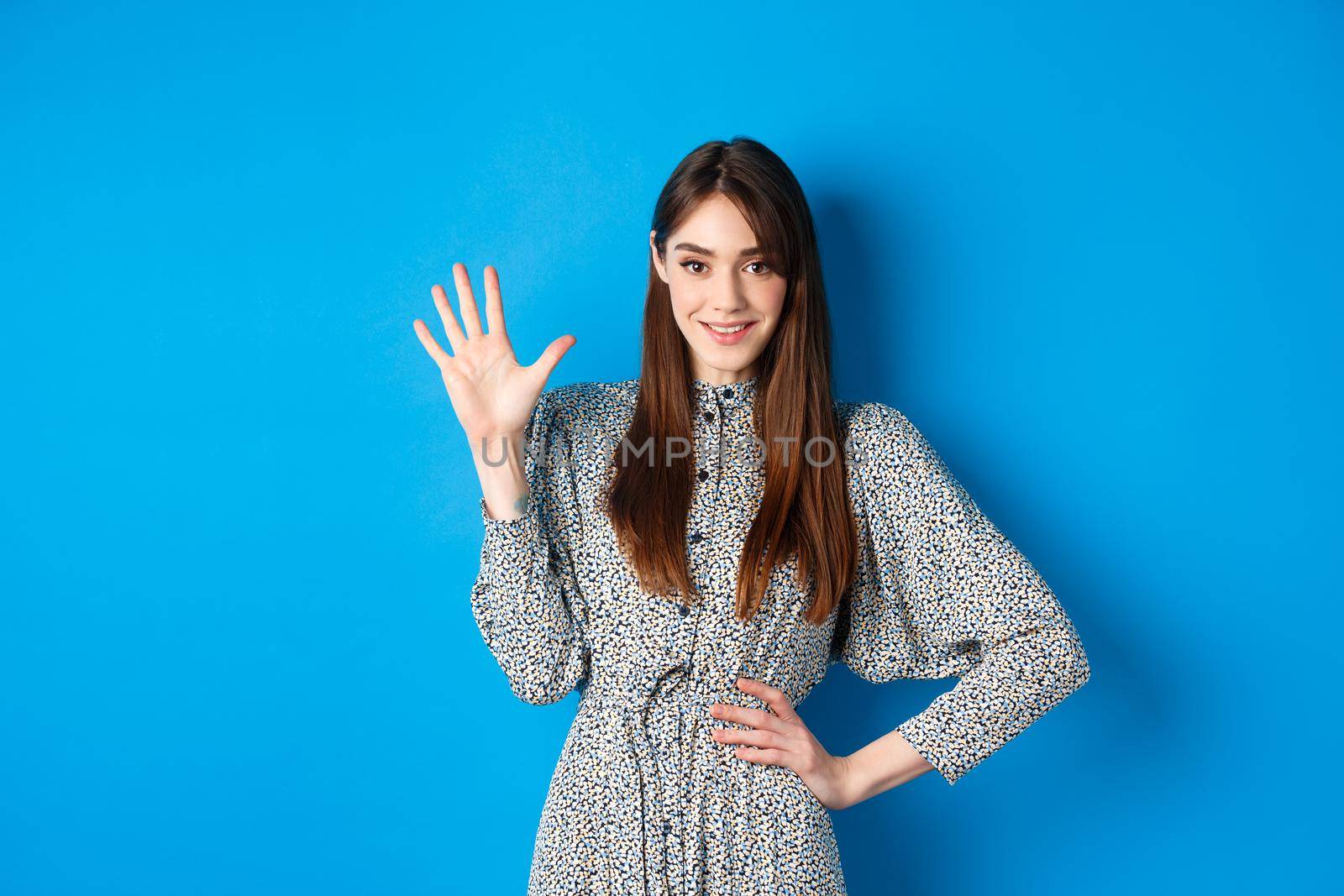 Young beautiful girl in dress with natural long hair, showing number five with fingers and smiling, standing against blue background.