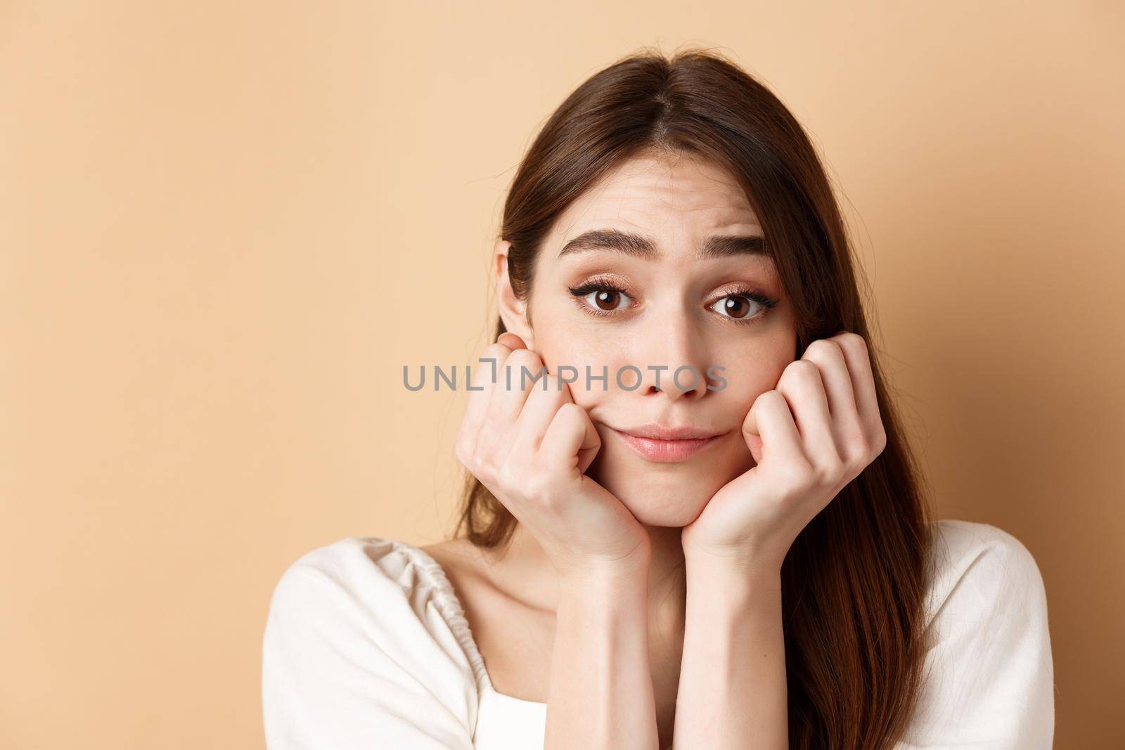Close-up of cute and pretty girl lean face on hands, looking at camera with dreamy smile, standing on beige background.