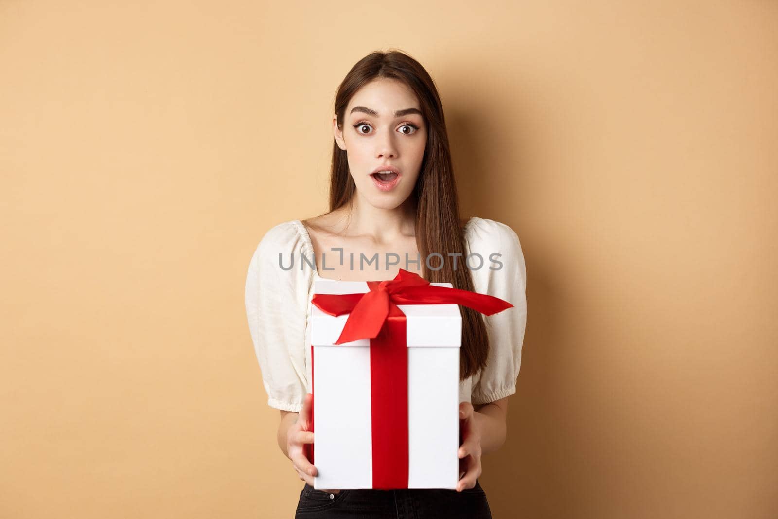 Amazed beautiful girl receiving gift from secret admirer on Valentines day, looking surprised at camera, holding box and standing on beige background.