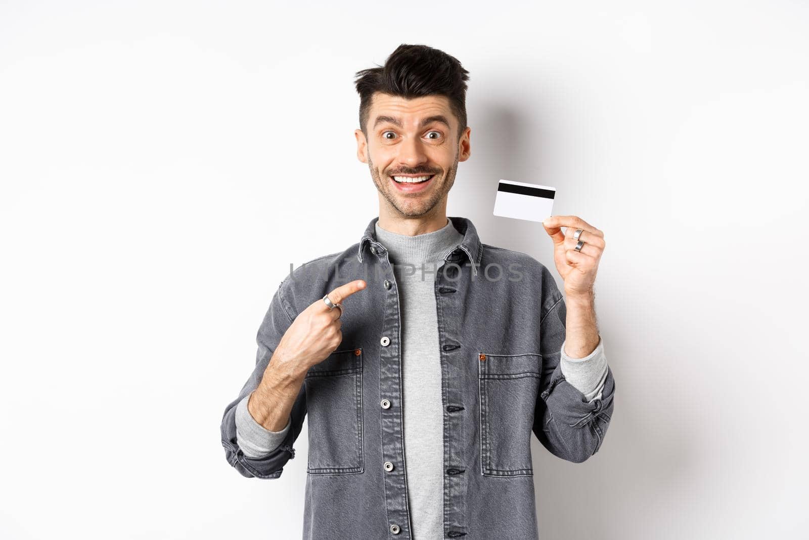 Excited handsome guy with moustache pointing finger at plastic credit card, smiling pleased, recommend good deal, standing on white background.