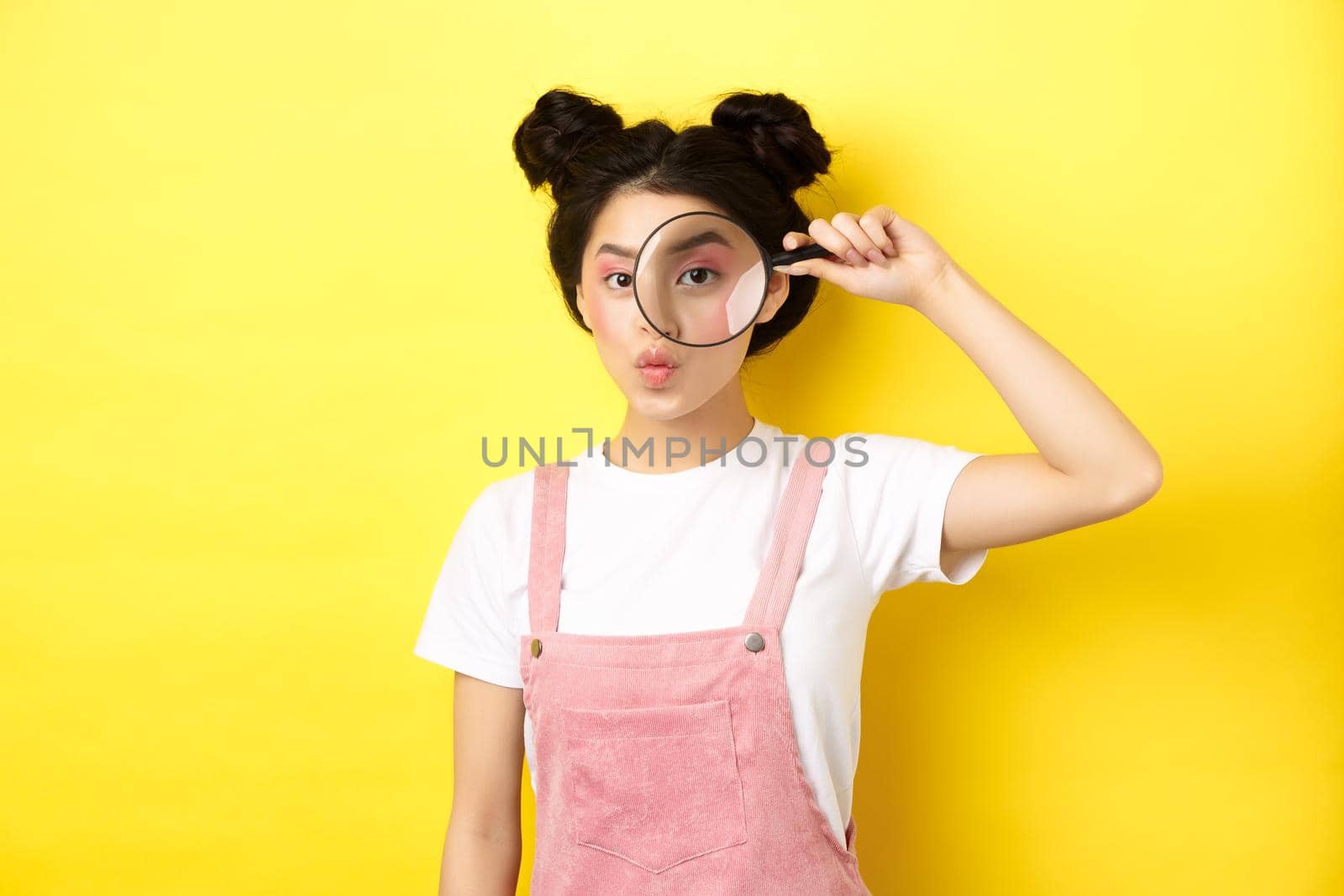 Silly glamour lady with bright makeup, look through magnifying glass and pouting, she is watching you, standing on yellow background.