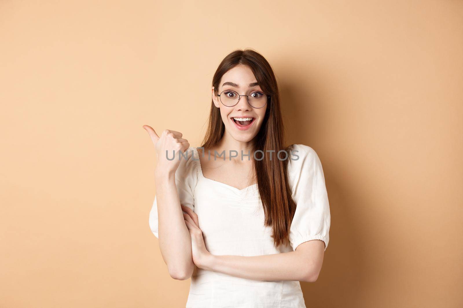 Excited girl in glasses pointing aside at logo, smiling amazed, recommending company, standing on beige background by Benzoix