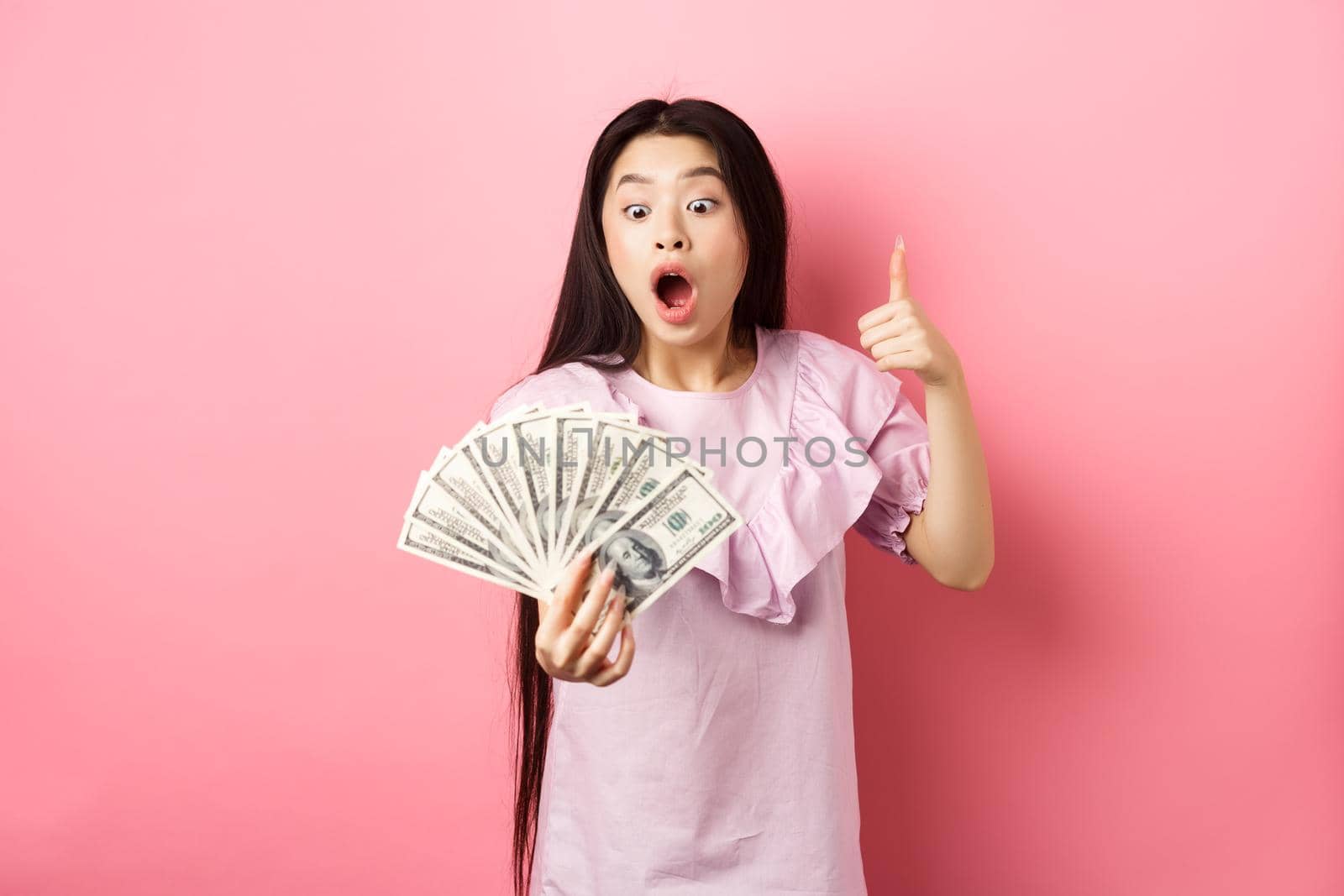 Excited teen girl holding big amount of money, showing dollar bills and thumbs up, standing amazed on pink background.