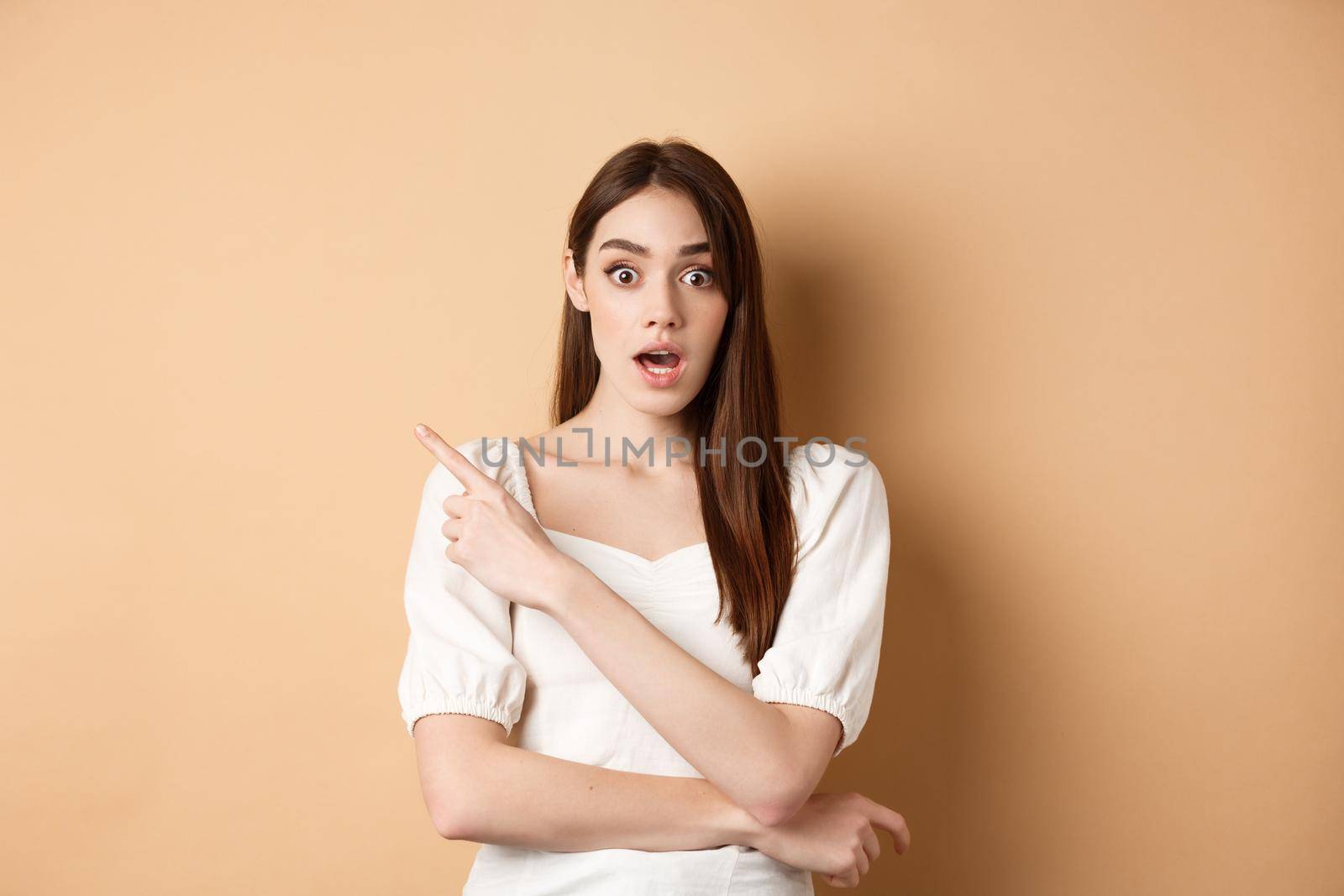 Surprised young girl say wow, pointing finger left at interesting banner, standing curious against beige background.