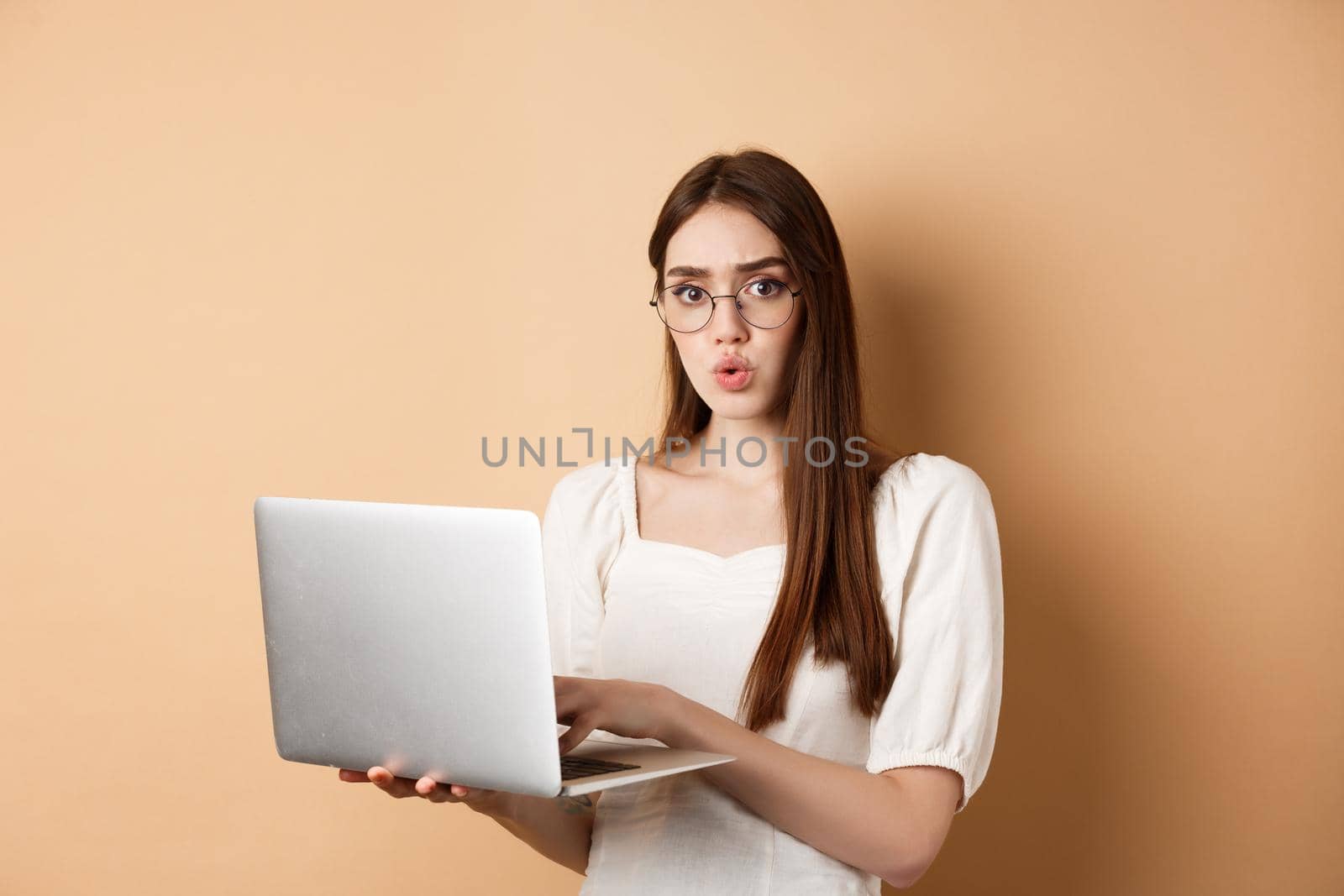 Concerned girl in glasses look at camera confused, working on laptop, having trouble with computer, standing on beige background.