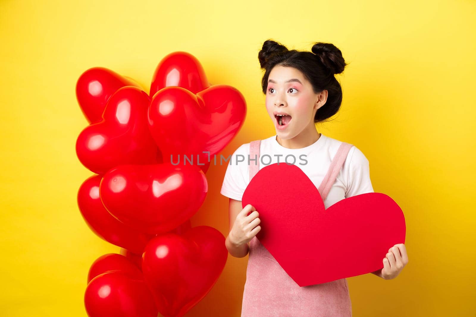 Happy Valentines day. Excited and surprised asian girl checking out romantic offer, looking left cheerful, showing big red heart card, yellow background.