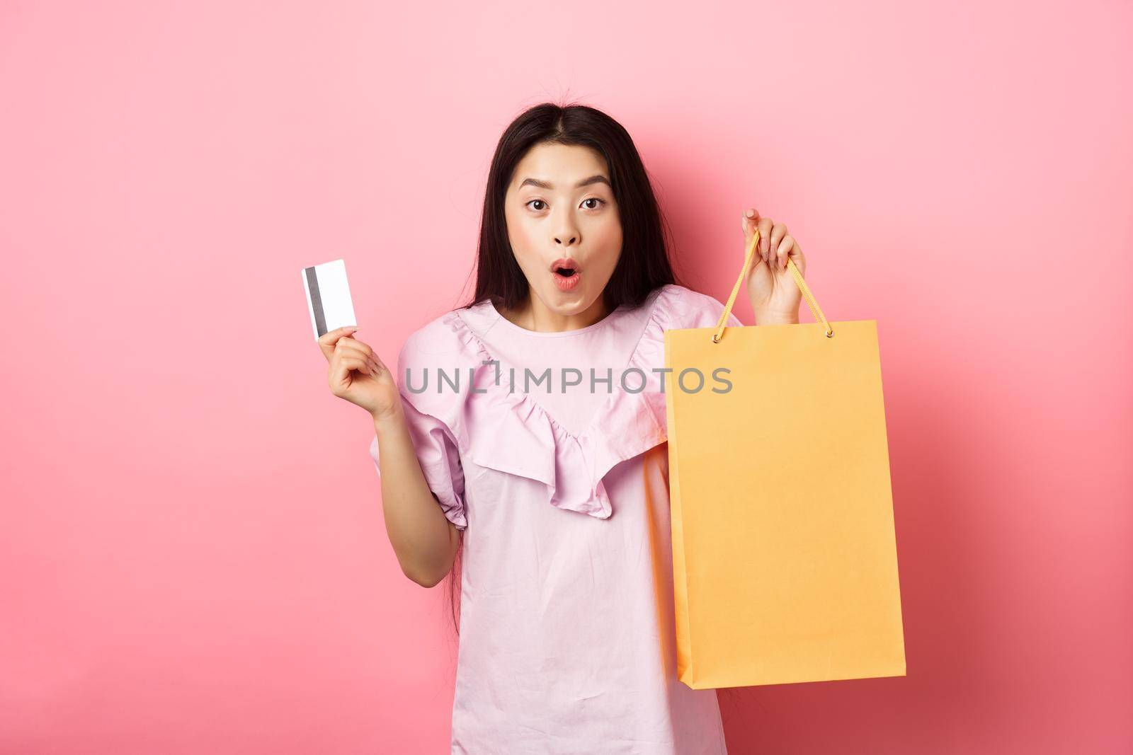 Shopping. Excited asian woman gasping amazed, holding bag from shop and plastic credit card, standing on pink background by Benzoix