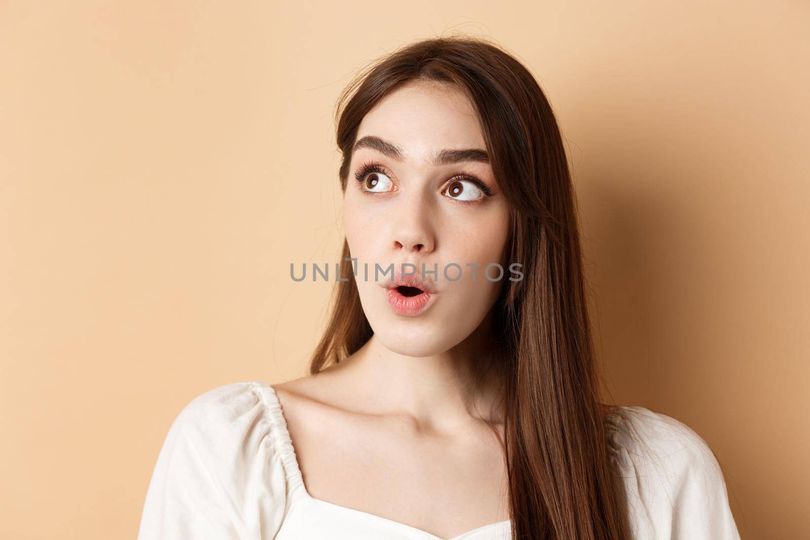 Close up portrait of excited pretty woman looking aside logo, say wow with amazed face, standing on beige background.