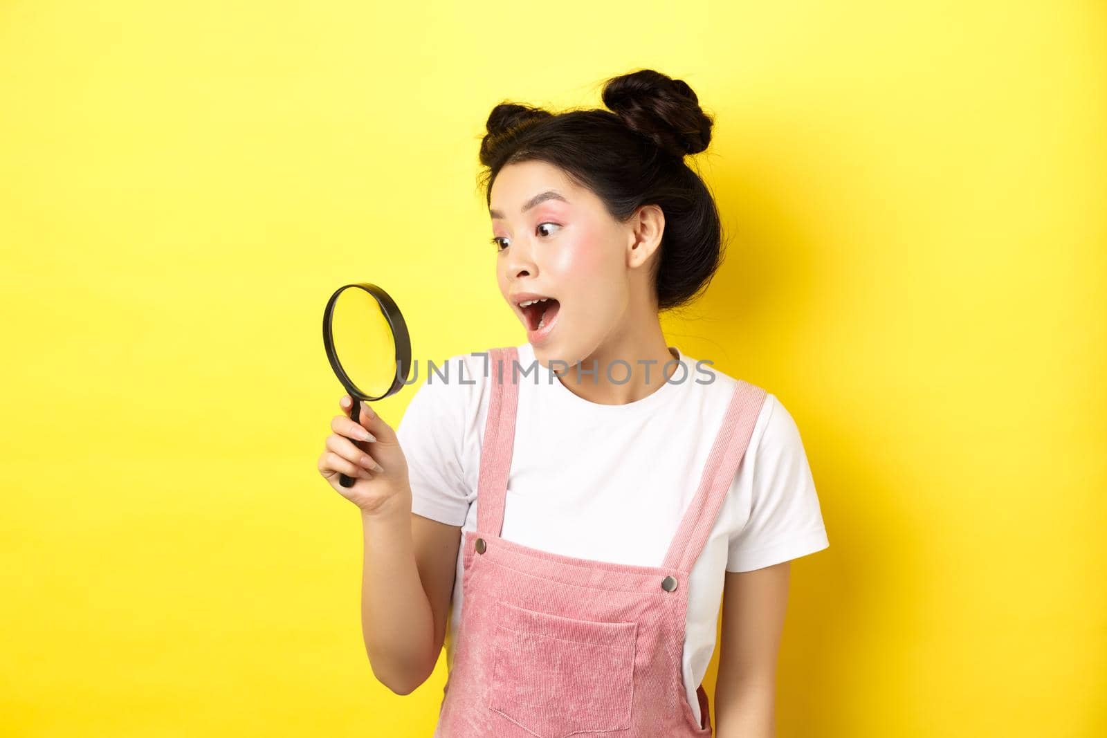 Excited asian girl found interesting thing, looking through magnifying glass amazed, standing on yellow summer background by Benzoix