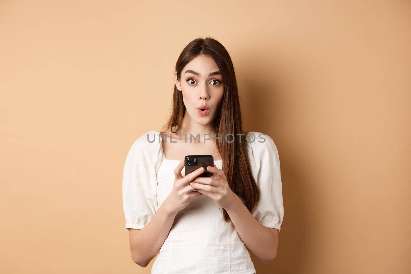 Girl standing amazed with smartphone, saying wow and look excited by awesome online offer news, standing on beige background by Benzoix