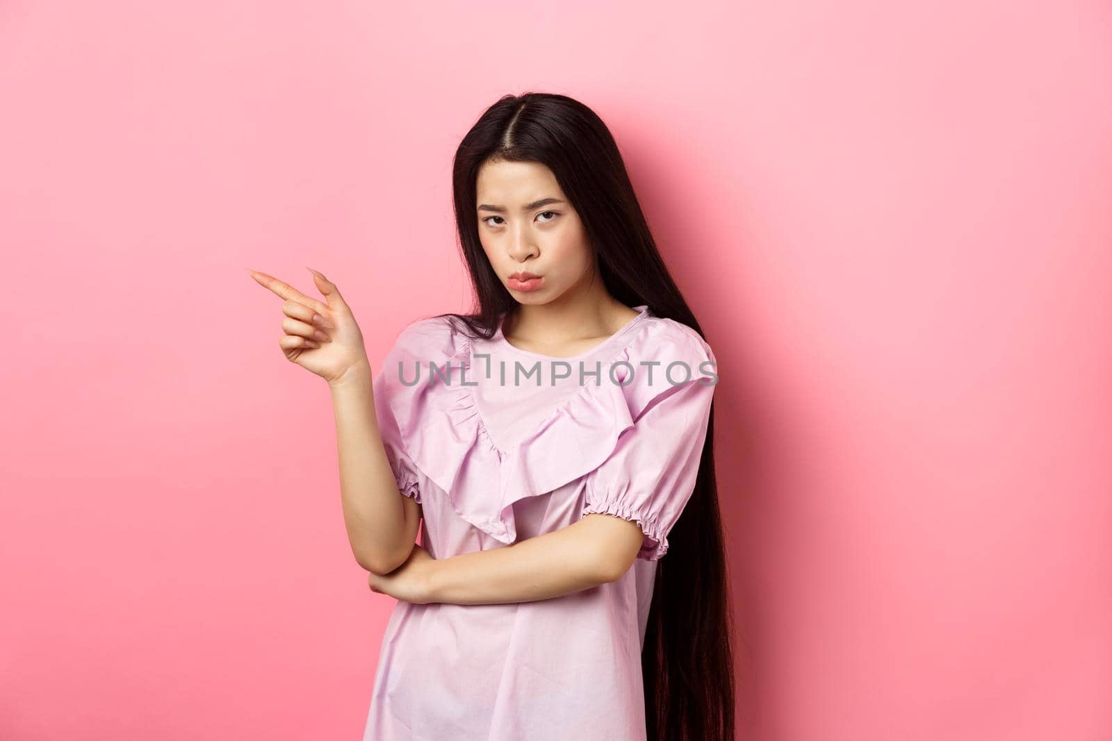 Sad and grumpy asian girl sulking, pointing left and look at camera jealous, want something, complaining and showing desired thing, standing on pink background by Benzoix