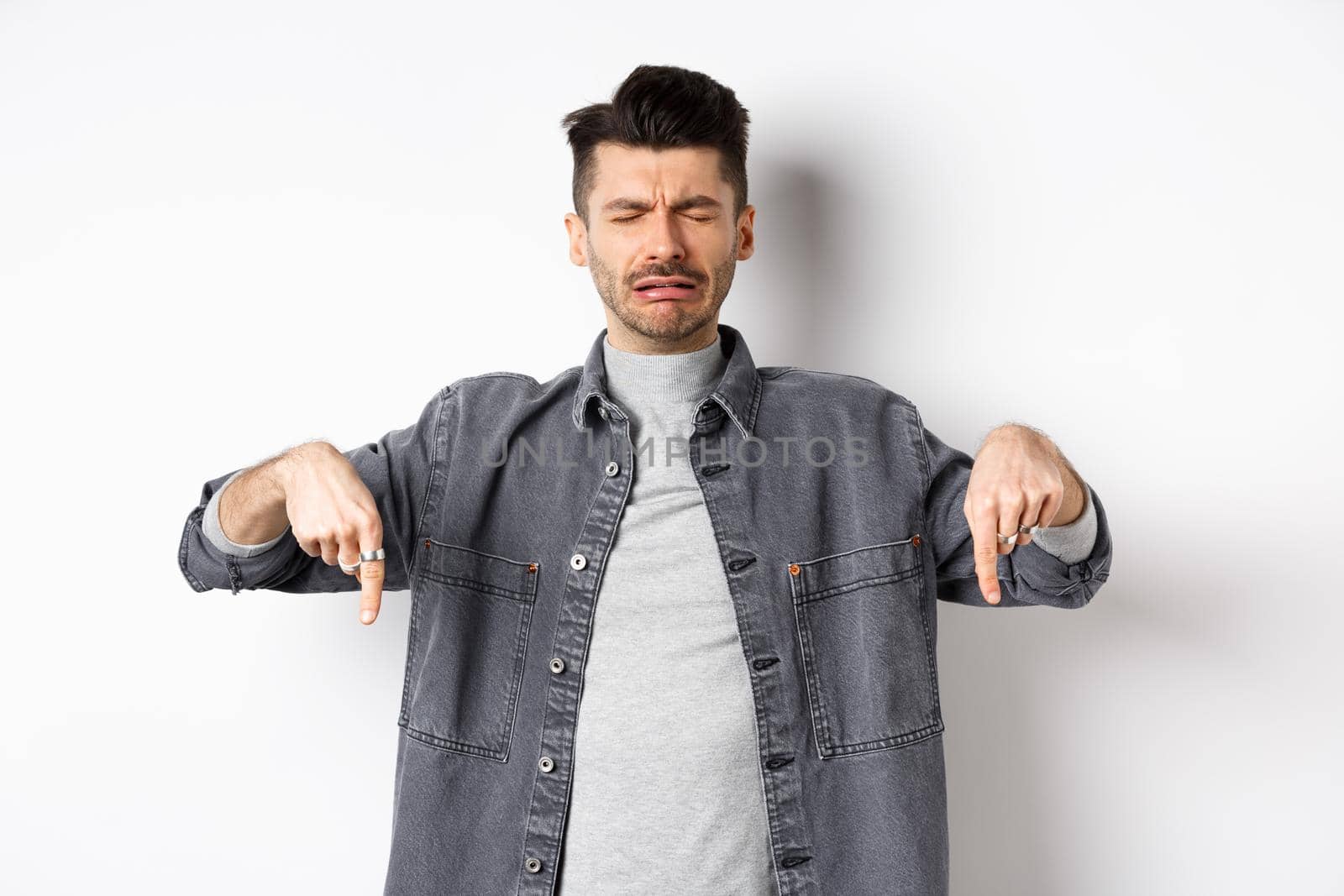Sad crying man whining, pointing fingers down disappointed, showing bad news, standing upset on white background.