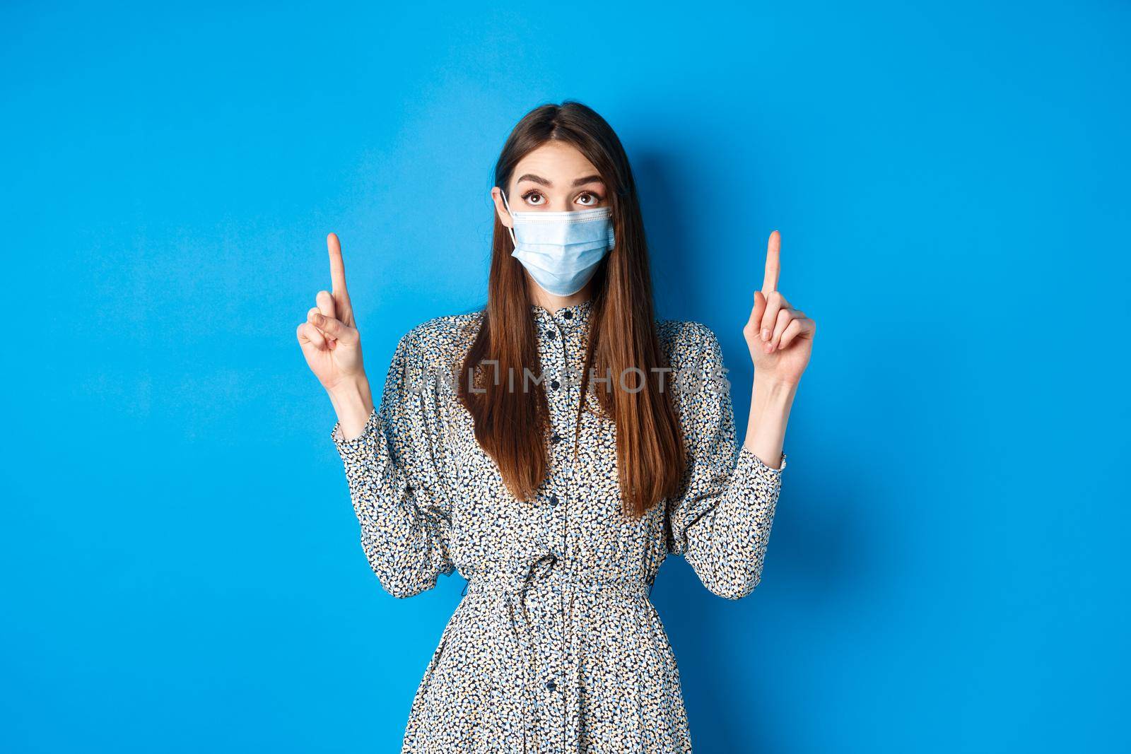 People, covid and quarantine concept. Intrigued young woman looking and pointing up at advertisement, standing in dress and face mask from coronavirus, blue background.