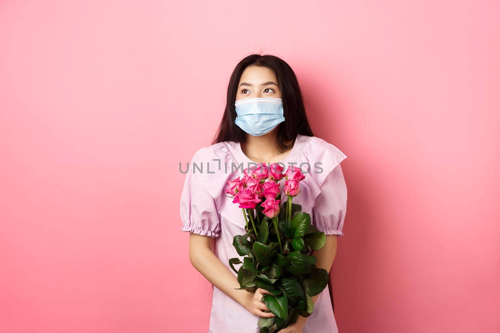 Romantic asian girl in medical mask looking aside at empty space with dreamy eyes, holding bouquet of roses for Valentines day, having date with lover during pandemic by Benzoix