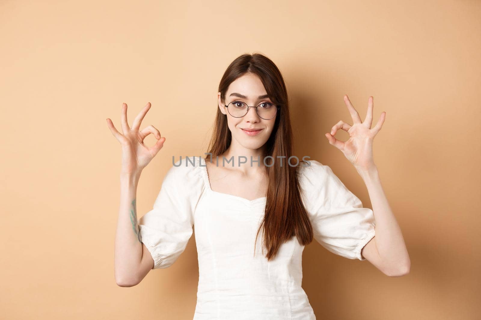 Satisfied female model in glasses show okay sign, looking pleased, agree and approve, standing on beige background.