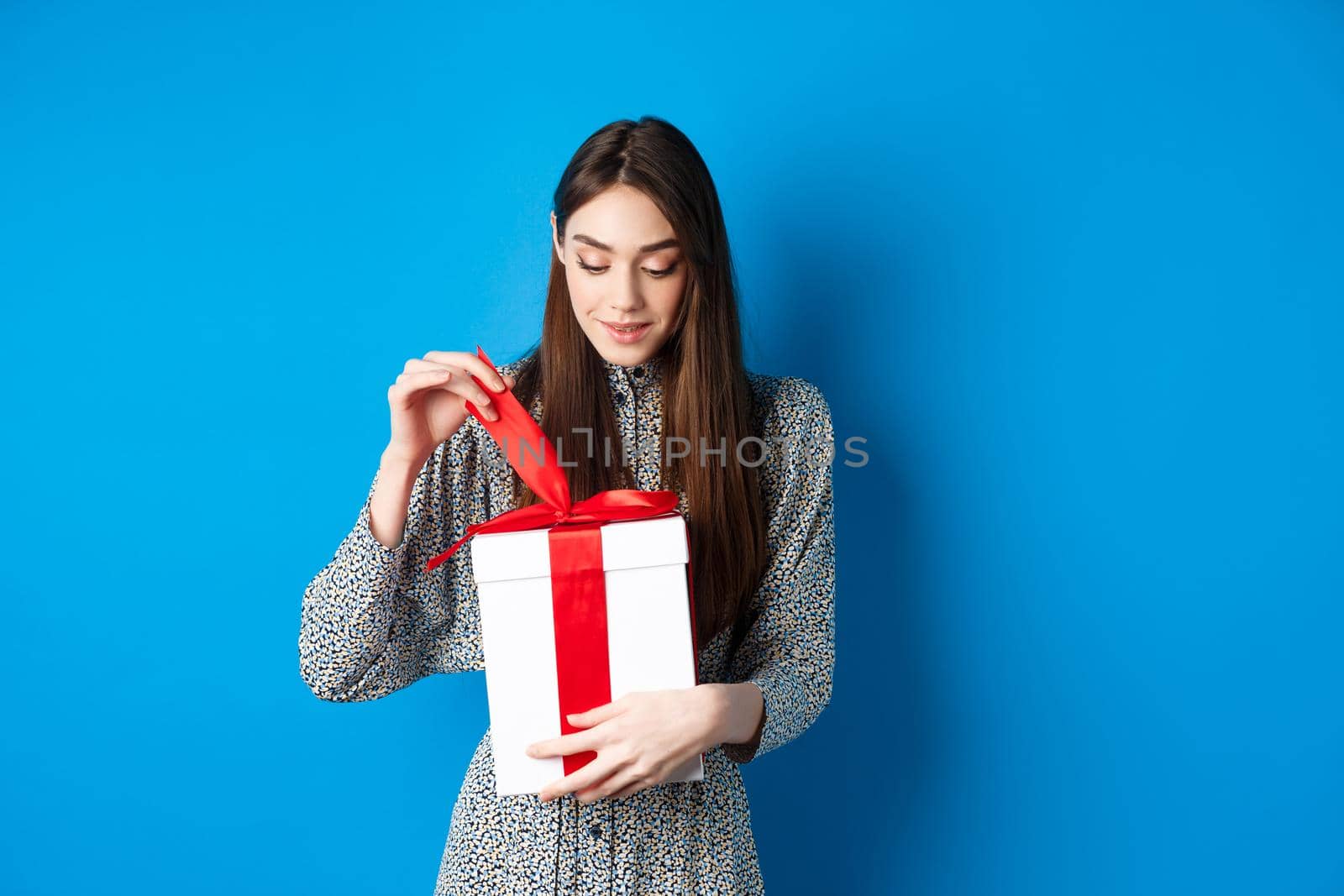 Valentines day. Cute young woman open box with gift, take-off ribbon from present and smiling intrigued, standing on blue background by Benzoix