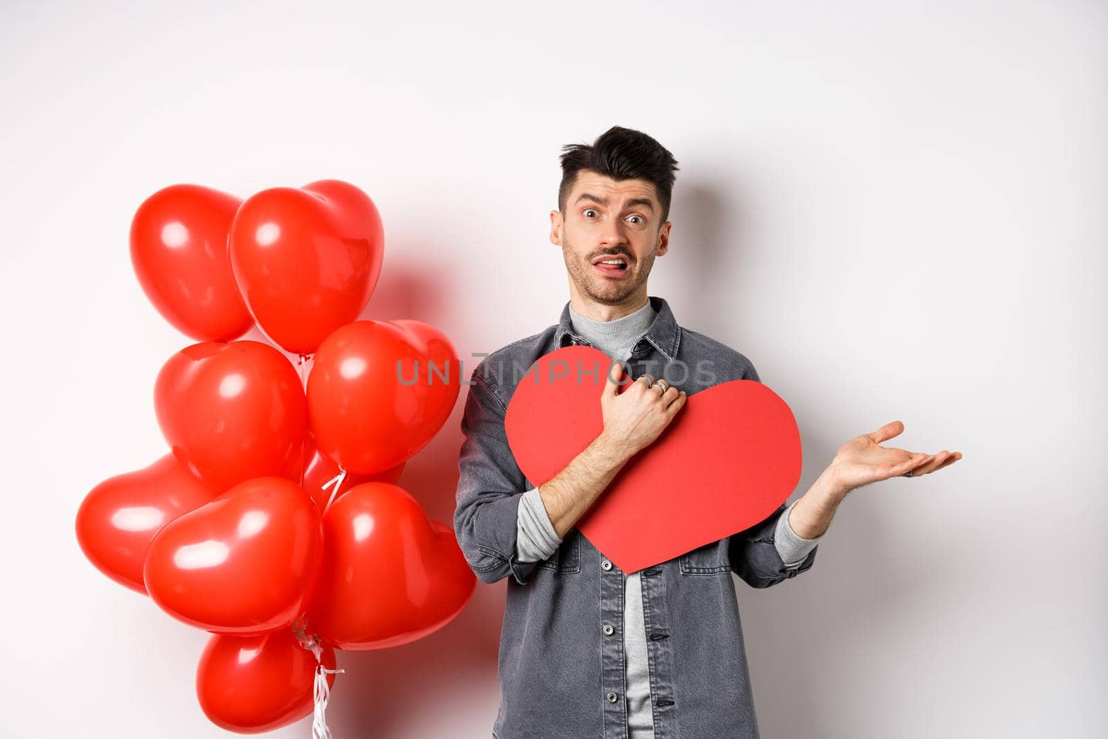 Confused man hugging big Valentine card and shrugging, feeling careless about romantic holiday, standing skeptical on white background by Benzoix