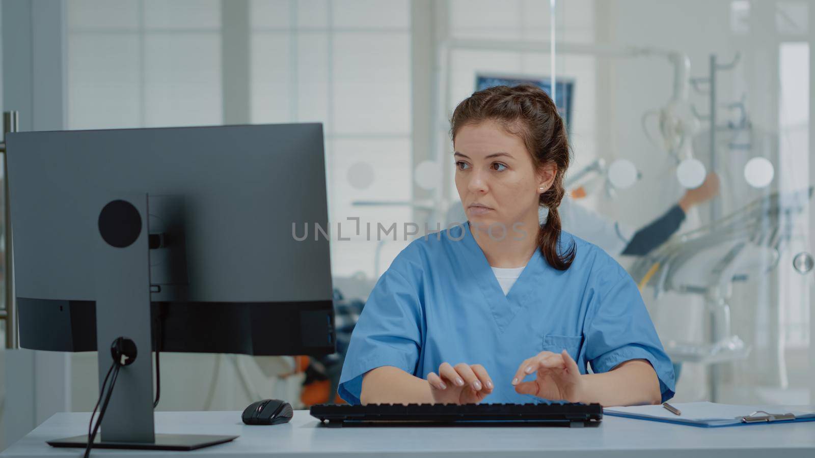 Stomatological assistant typing on computer keyboard sitting at oral clinic desk. Nurse using technology while professional orthodontist consulting patient with equipment in background