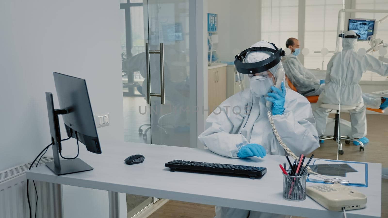 Dentistry nurse in protection suit sitting at desk using telephone and computer at dental clinic. Assistant working on appointments while stomatologist consulting patient in background