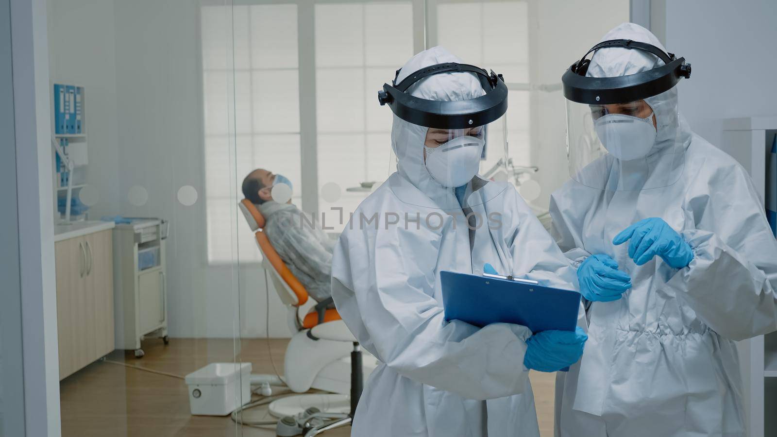 Professional stomatologists wearing ppe suits at oral clinic discussing dental operation for patient sitting in cabinet. Dentists preparing for consultation during coronavirus pandemic