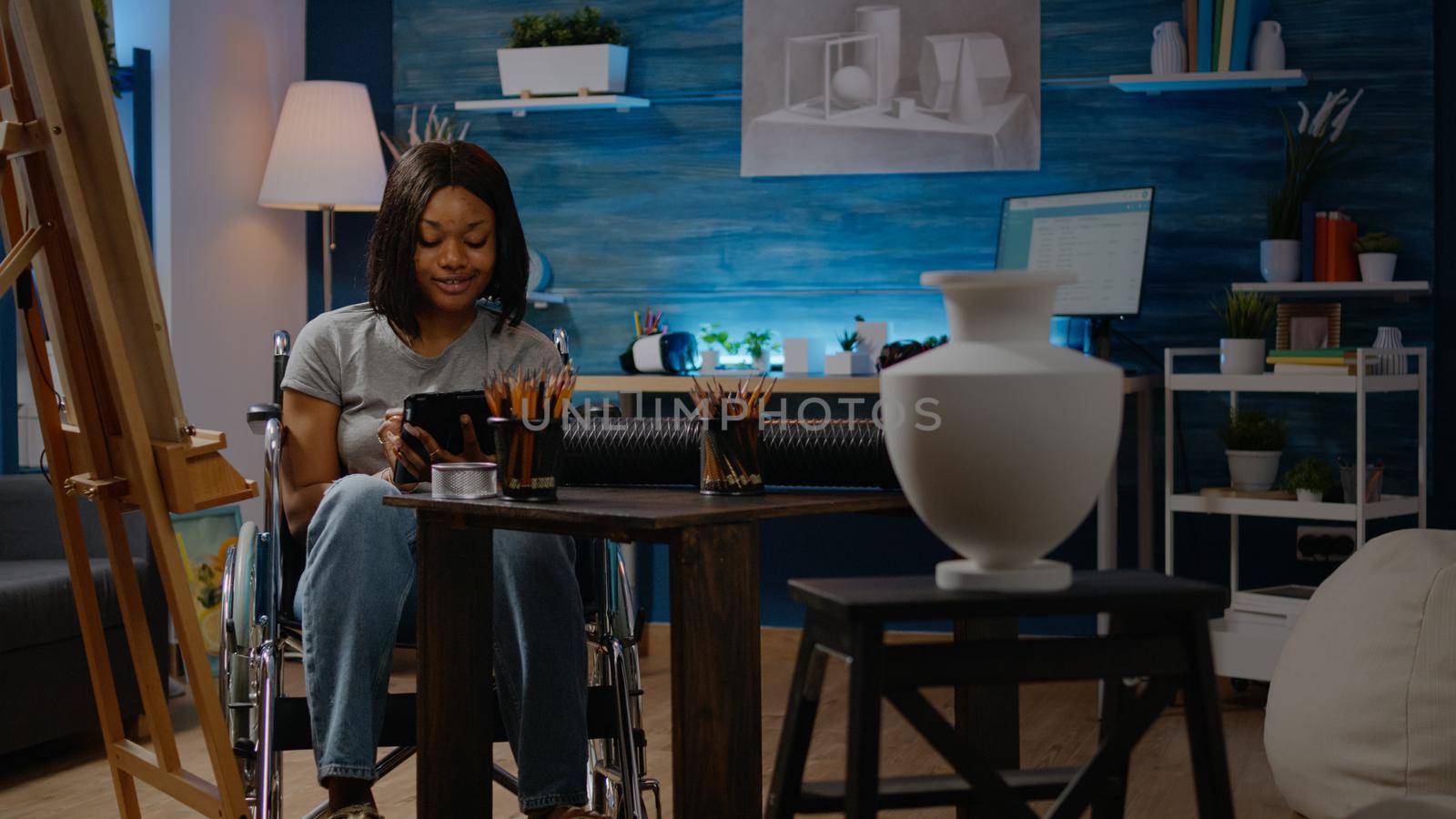 African american woman with disability holding digital tablet working on vase drawing in studio space. Black artist in wheelchair using technology for masterpiece design on canvas