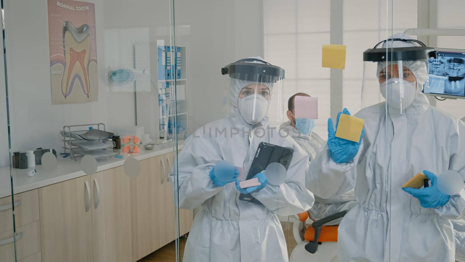 Dentistry team of specialists using sticky notes on glass in stomatology cabinet to explain teethcare diagnosis to patient. Dentist and nurse wearing ppe suits for examination during pandemic