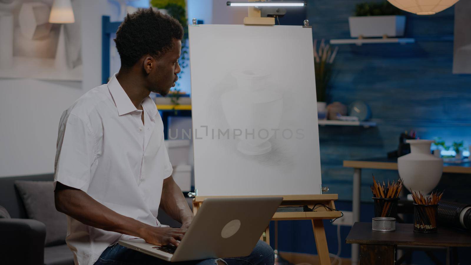 Black creative artist holding laptop computer in art studio space. Young man of african american ethnicity using technology for artistic drawing of vase as professional masterpiece