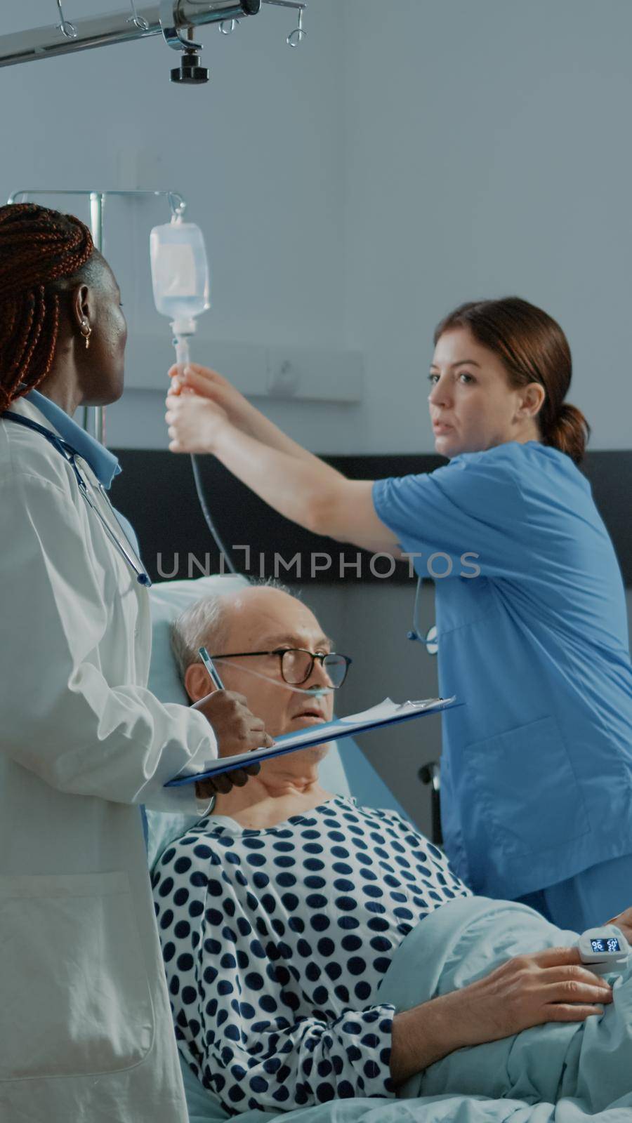 Nurse and african american doctor treat sick patient in hospital ward with modern medical equipment. Old man with nasal oxygen tube and oximeter waiting for treatment against disease