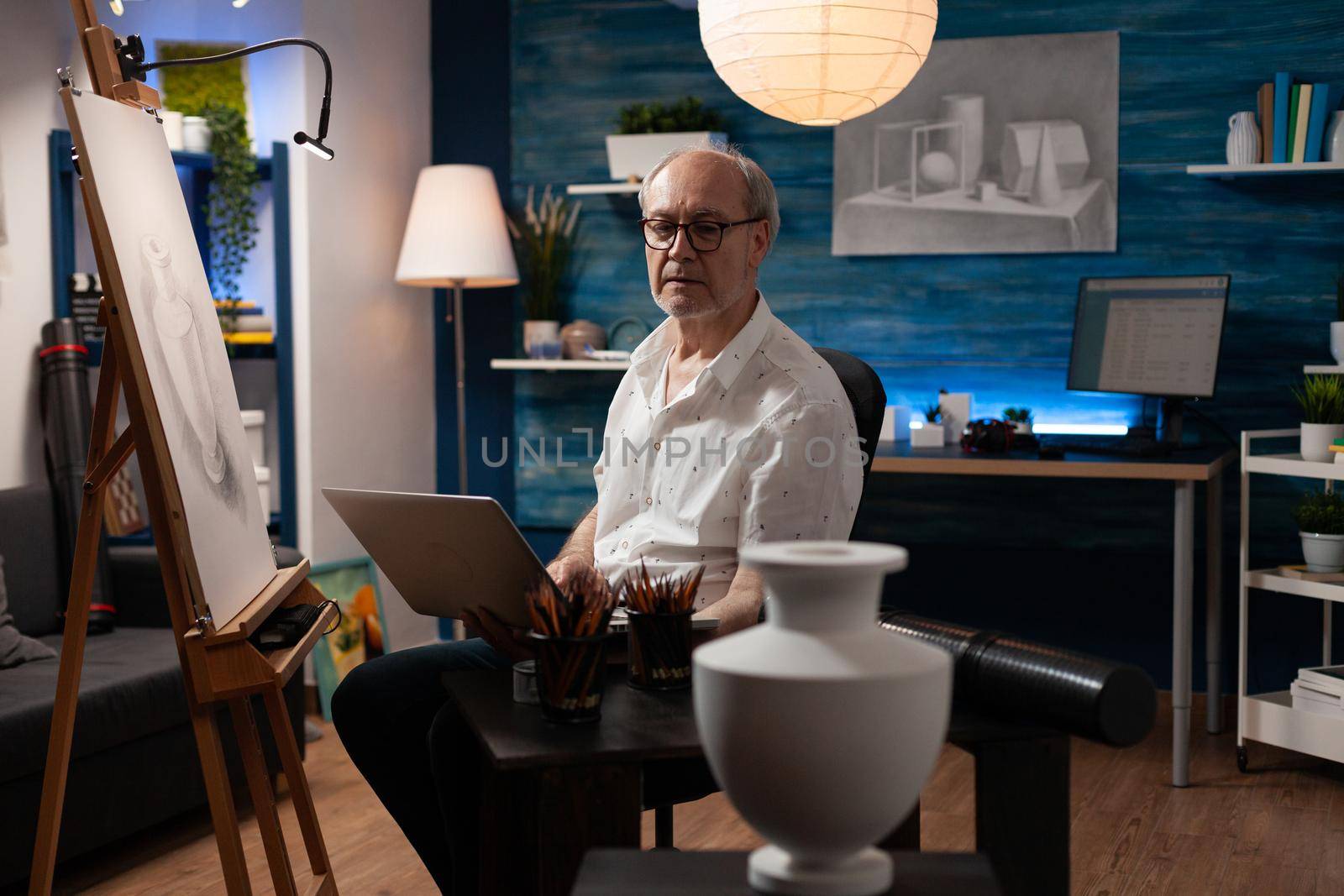 Senior artist holding laptop for drawing creation of white vase on table at art studio. Elderly person with imagination working on authentic masterpiece and design on canvas with easel