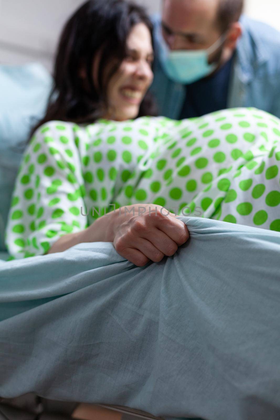 Close up of hand grabbing bedsheet from painful labor contractions at maternity. Young woman giving birth to child while father of baby supporting wife in hospital ward background