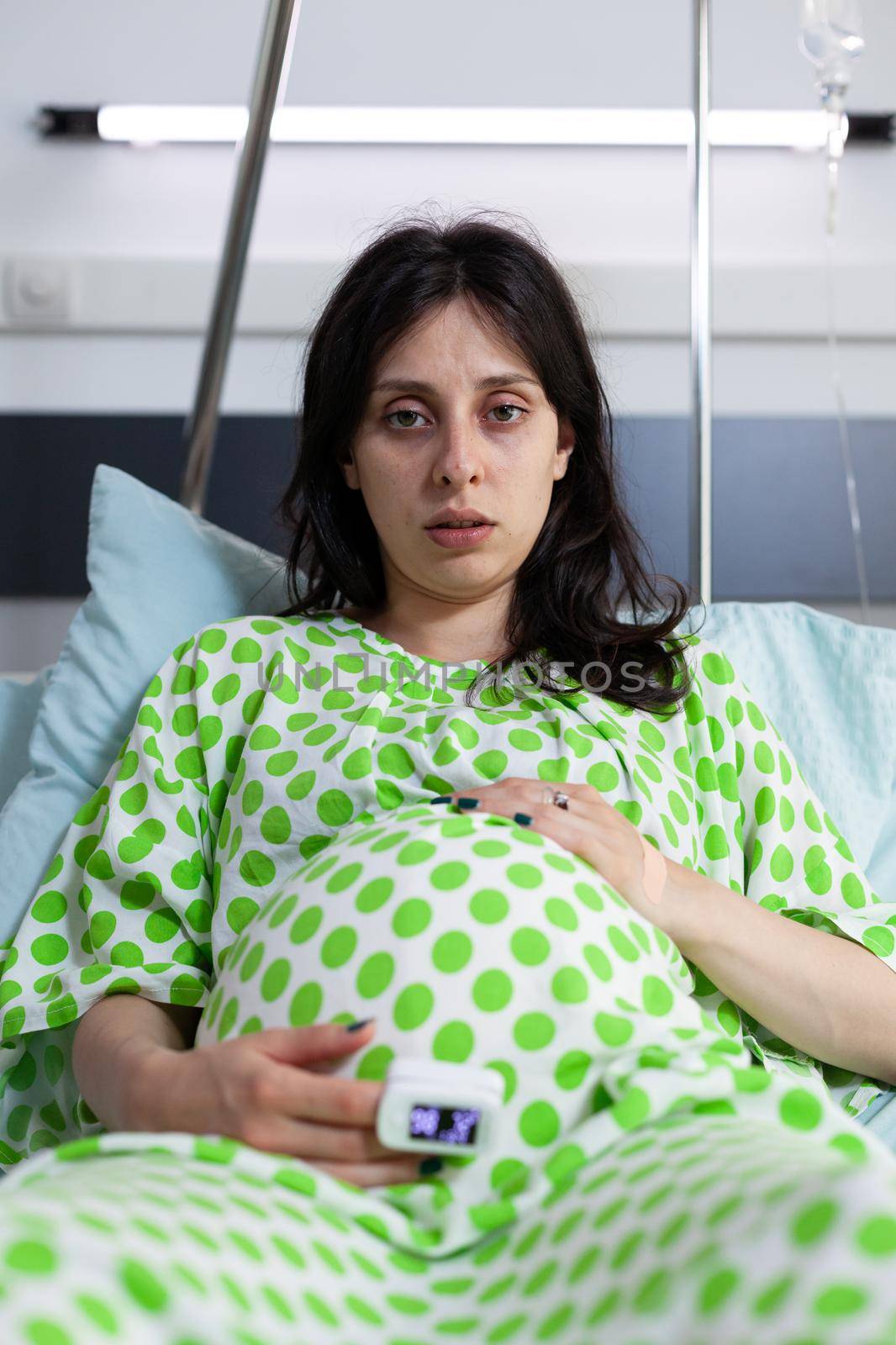 Portrait of tired pregnant person sitting in hospital ward bed by DCStudio