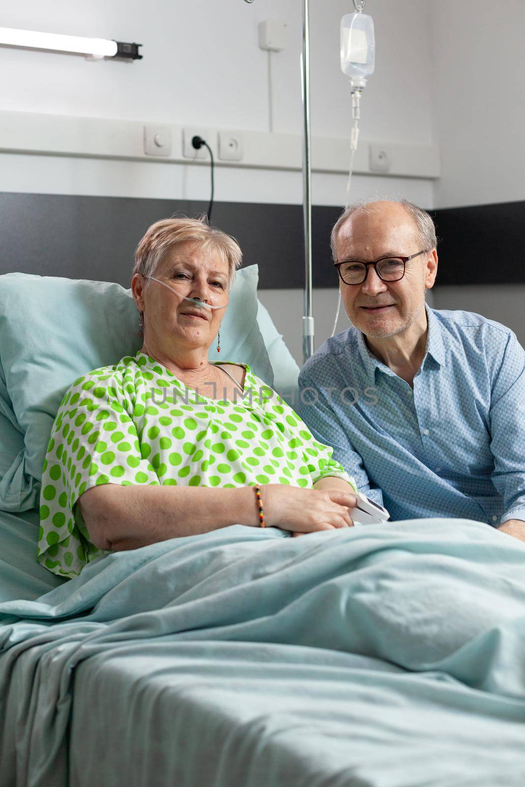 Retired hospitalized sick woman lying in bed with husband visitor sitting beside her waiting together for medical treatment. Senior man visiting patient during medicine examination in hospital ward