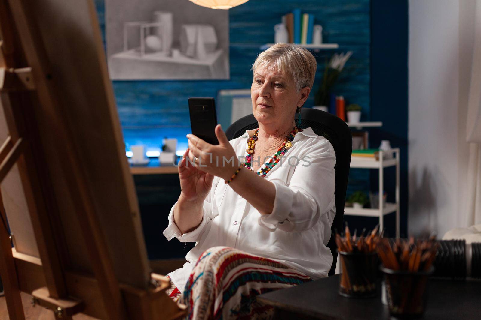 Senior artist using smartphone sitting in art studio room by DCStudio