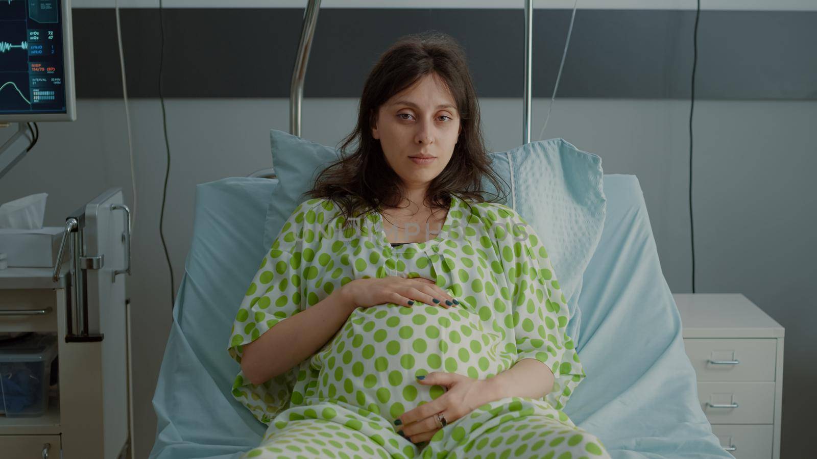 Portrait of pregnant woman laying in hospital ward bed with medical equipment preparing for childbirth at maternity. Caucasian patient holding hand on belly while sitting expecting baby