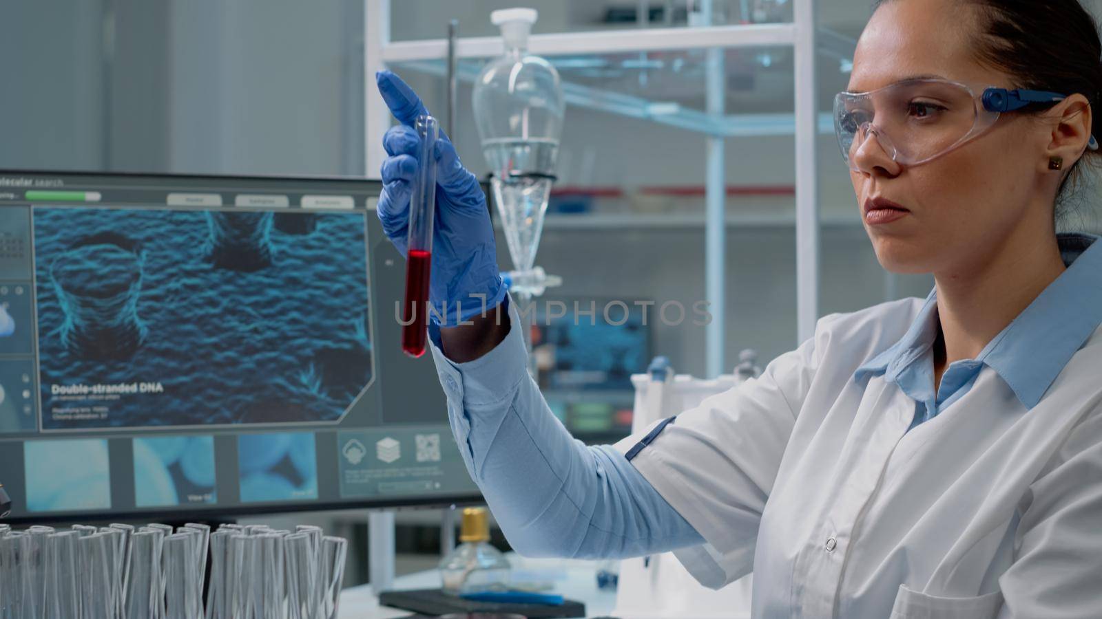 Medicine scientist using computer while holding test tube with red liquid solution for research development. Biochemistry woman with protection glasses and gloves studying lab flask