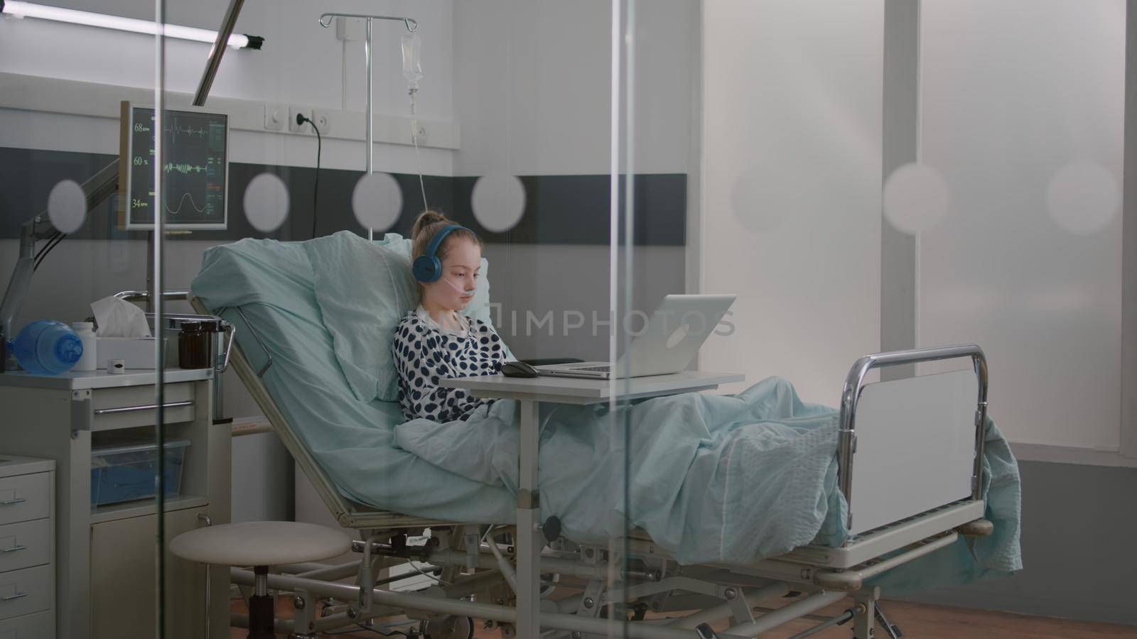 Alone sick girl patient with oxygen nasal tube relaxing in bed wearing headphones while watching entertainment movie on laptop computer during medical examination. Girl recovery after surgery