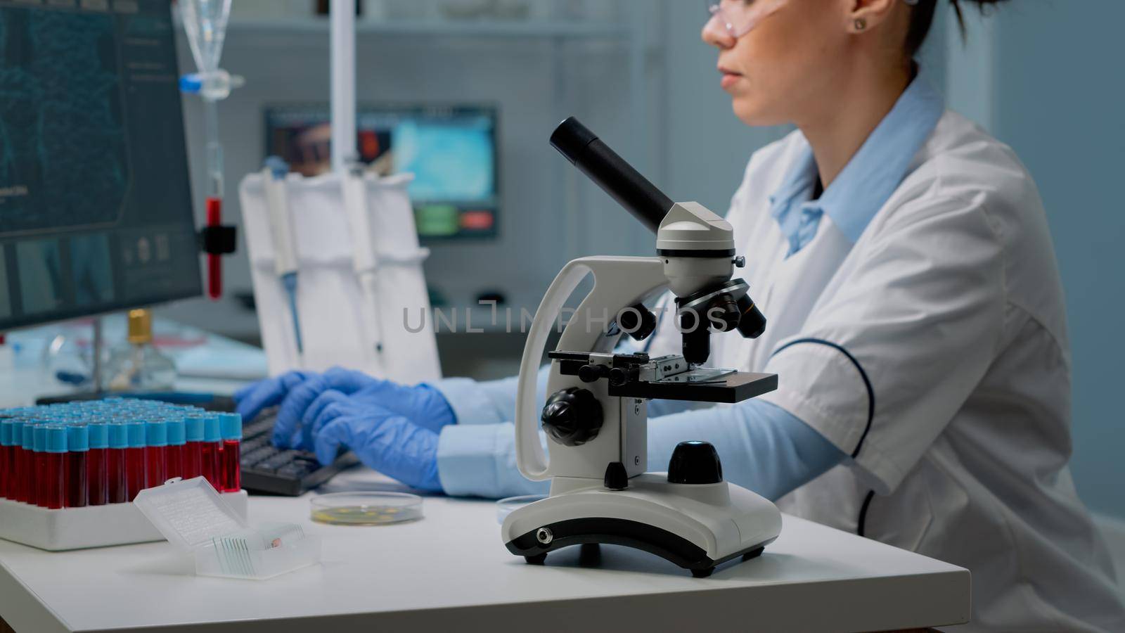 Close up of microscope tool while scientist analyzing petri dish and using modern computer in background. Woman at laboratory desk with blood sample and tray of vacutainers for examination