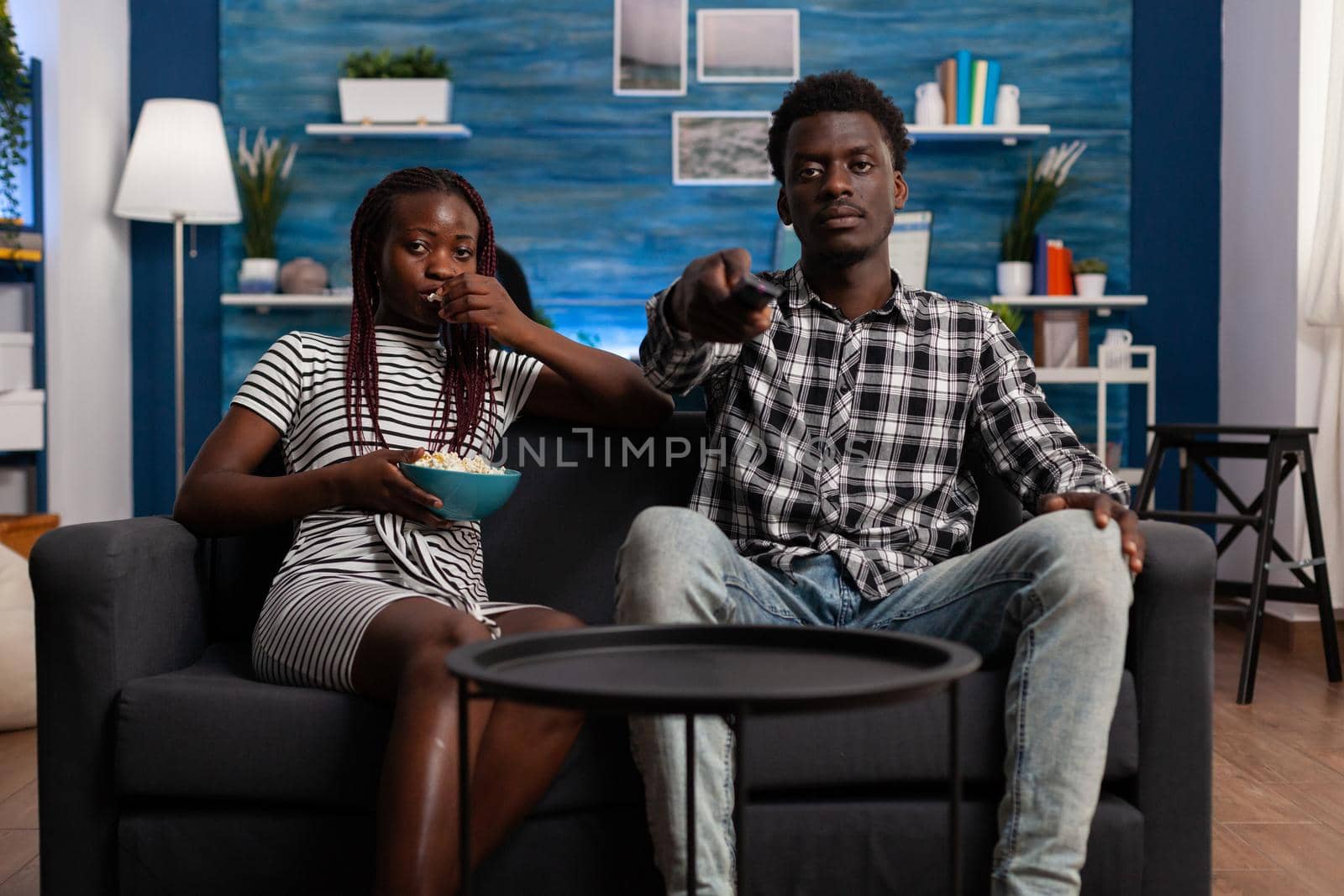 POV of black married couple on couch watching television and looking at camera. African american woman eating popcorn from bowl while man holding TV remote control at home