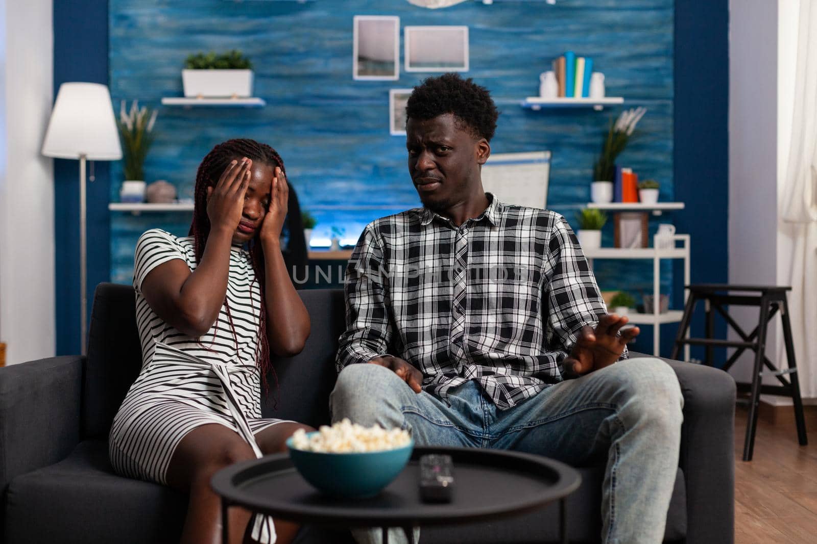 African american people watching scary movie on television and being afraid on sofa. Black woman covering eyes at TV while man looking at camera. Couple enjoying horror movie experience