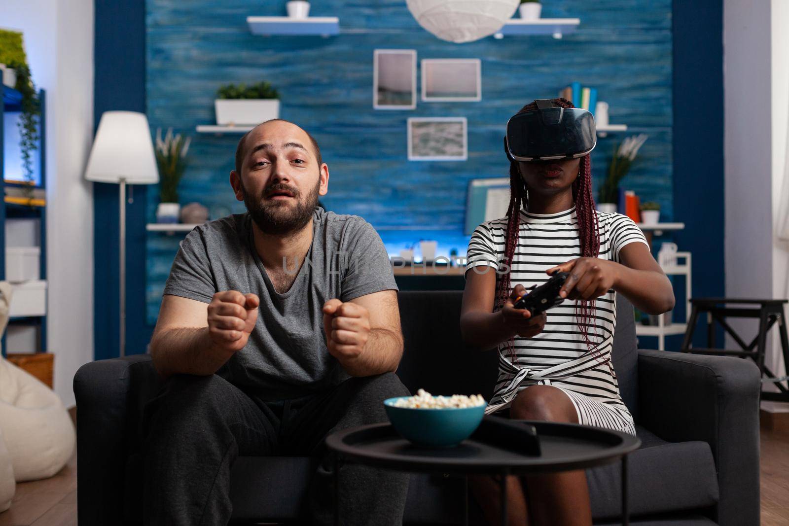 Interracial couple using vr glasses and controllers for fun at home. Mixed race young people with technology playing video game with goggles, headset and console on television.