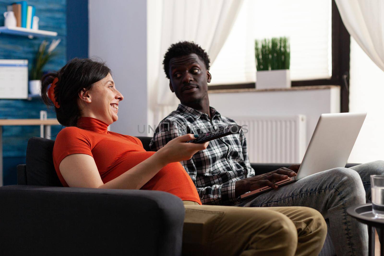 Interracial couple with pregnancy smiling and relaxing at home. Caucasian woman with baby bump holding remote control for television and african american father of child using laptop