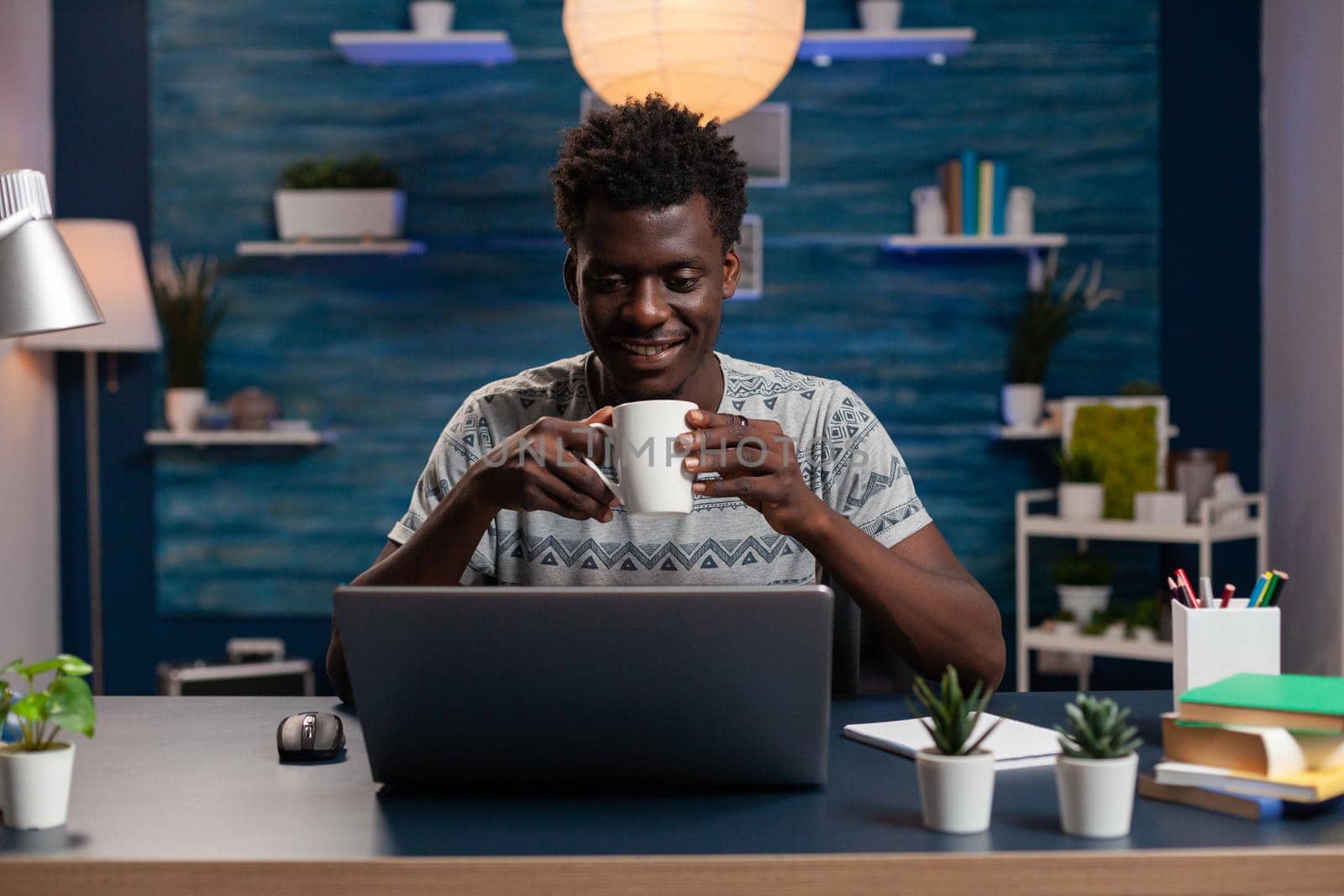 African american businessman holding cup of coffee typing management ideas on computer working remote at online project in living room. Freelancer sitting at desk analyzing financial graph