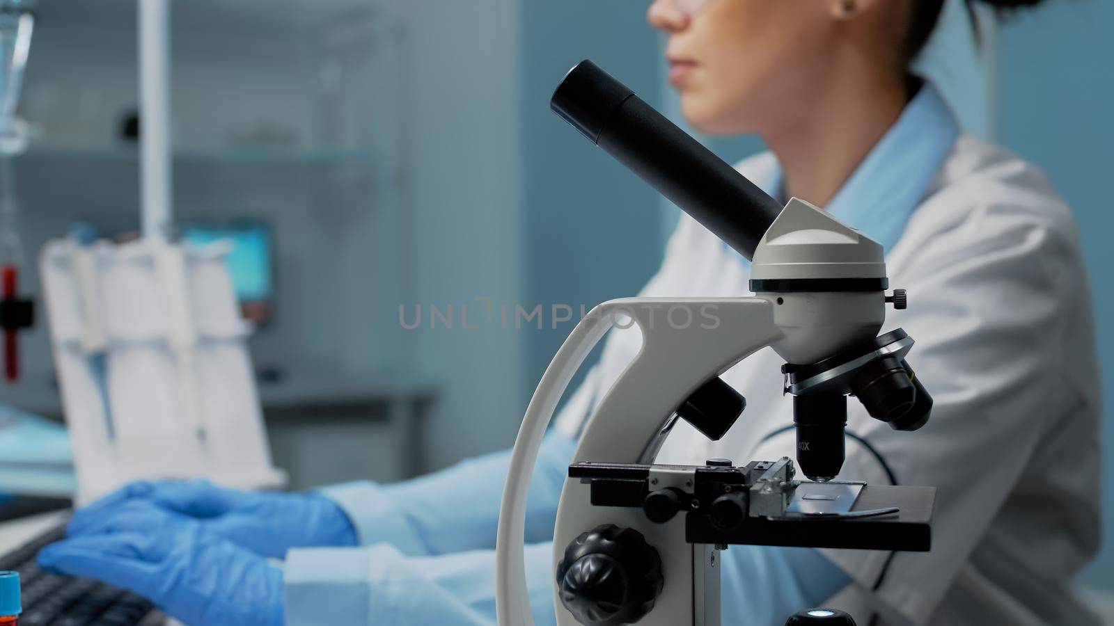 Scientific woman with microscope tool studying petri dish while using computer in background. Biology doctor in laboratory with research equipment and instruments for science innovation