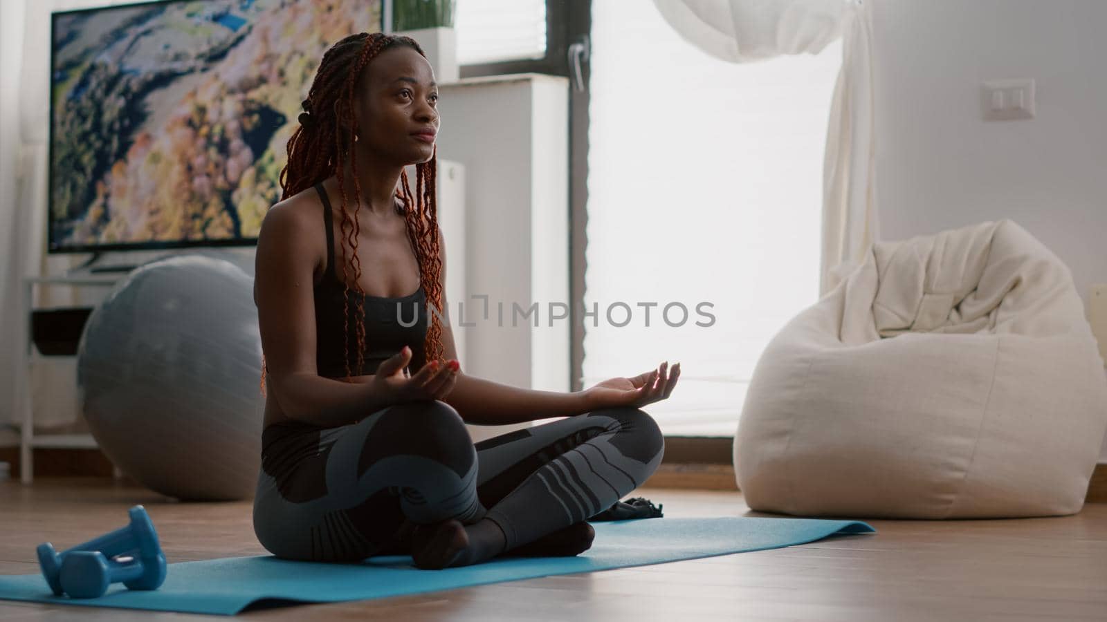 Flexible black athlete relaxing in lotus position on floor in living room by DCStudio