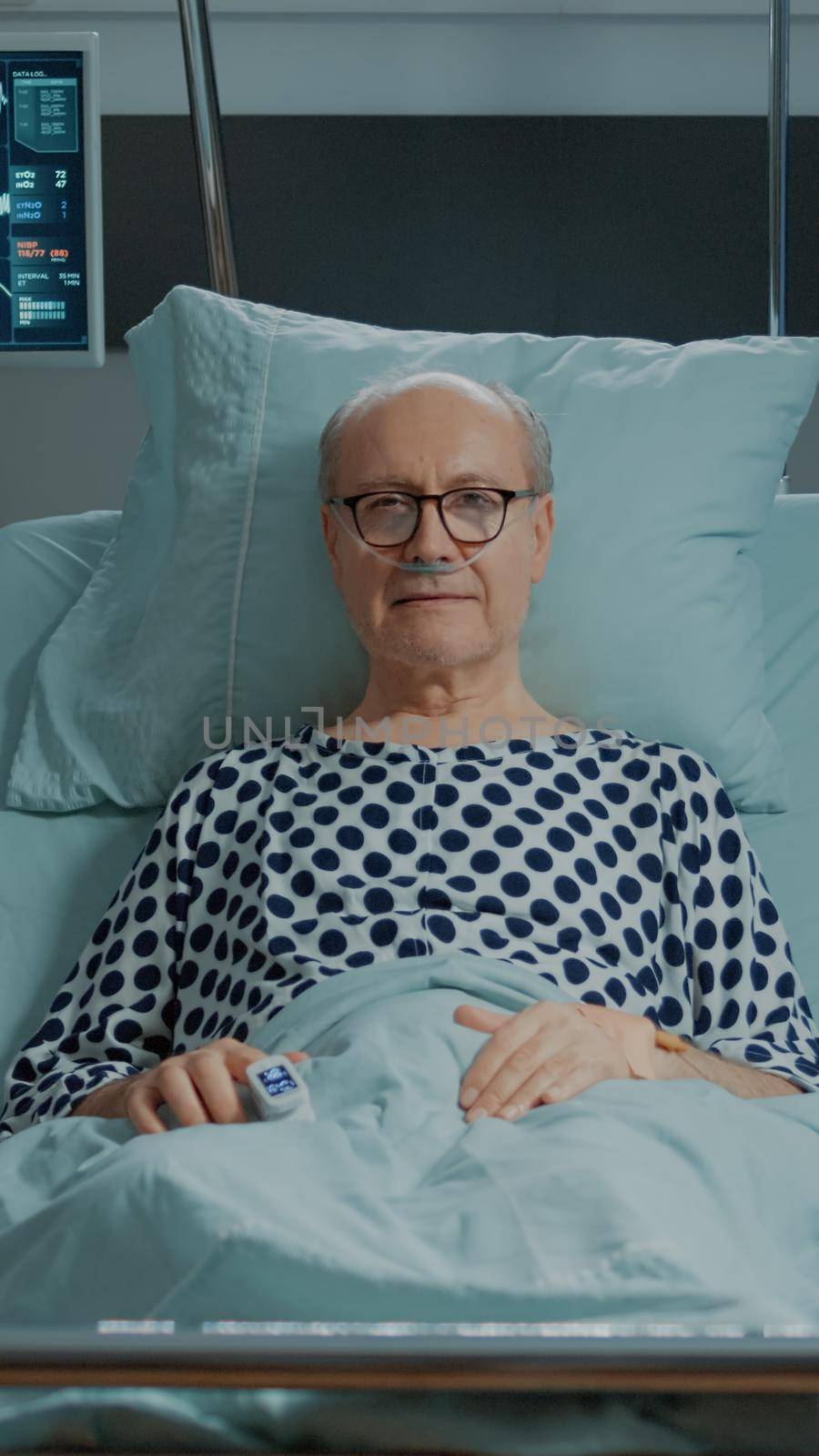 Portrait of sick patient staying in bed at hospital ward with oximeter, nasal oxygen tube and monitor. Old man waiting on doctor treatment to cure disease, illness, pain surgery and injury