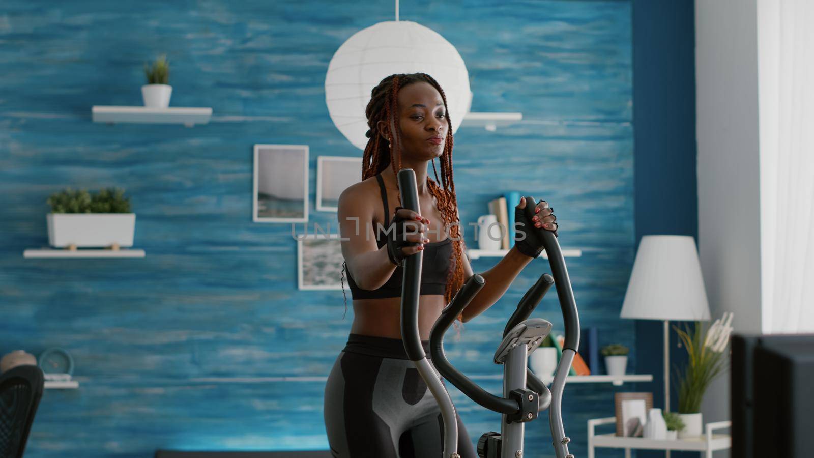 Fit athletic black woman running on elliptical bike watching wellness routine fitness video on tv during gym morning workout in living room. Athletic black woman training body muscle