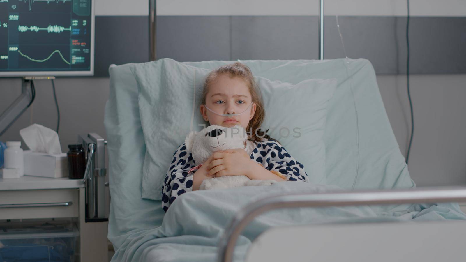 Portrait of lonley sick little child resting in bed holding teedy bear in hands looking at camera during recovery examination in hospital ward. Hospitalized kid waiting for healthcare treatment