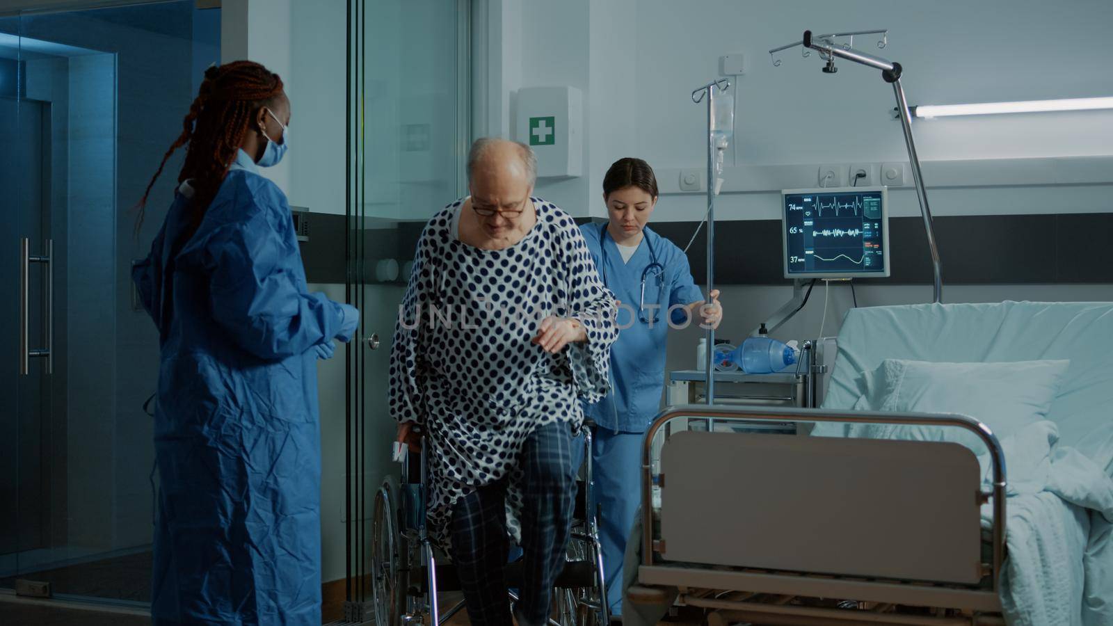 Nurse and african american doctor helping seat sick patient in hospital ward wheelchair at medical unit. Old man with IV drip bag preparing for modern technology surgery to treat injury