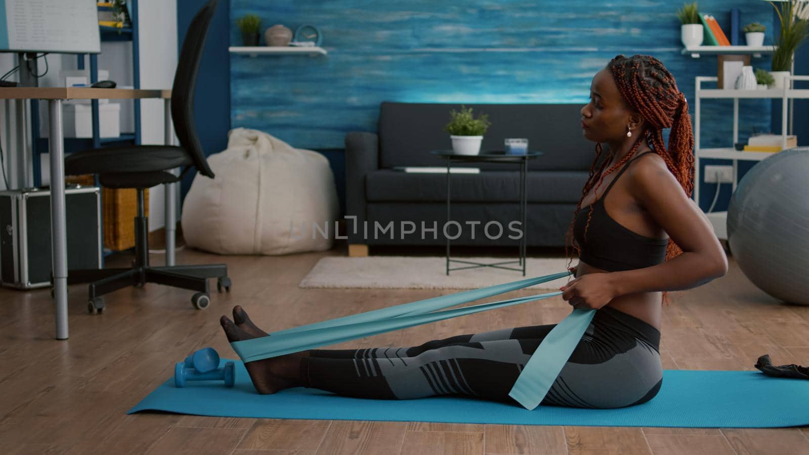 Slim woman with black skin in sportswear exercising body muscle using fitness bands practicing sport on yoga map in living room. Fit woman working at wellness warming before workout